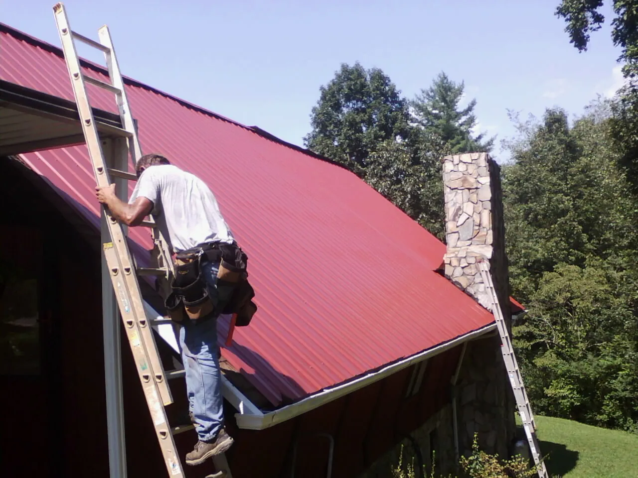 Roofer on a ladder beside a red metal roof and stone chimney — Soffit & Fascia Repair by Roofing Brothers in Michigantown